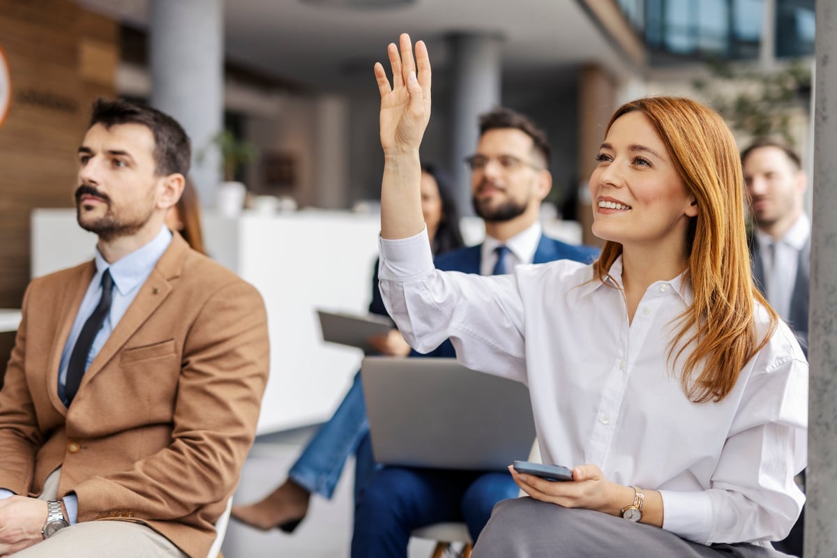 Female manager sitting with her team in a boardroom seminar, raising her hand to ask a question.